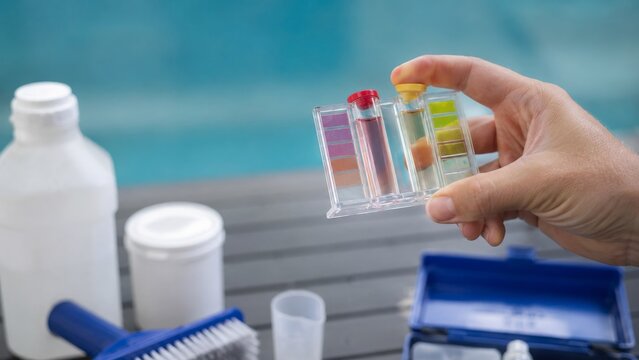 Hand holding pool water test kit with color comparator vials near bottles and blue case, testing water quality beside turquoise water outdoors