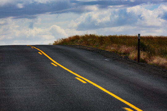 A two lane road with yellow central line, one solid and one broken through countryside.