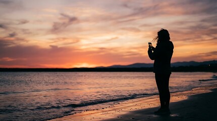 Fototapeta premium Silhouette of a woman taking a photo with her smartphone on a beach at sunset