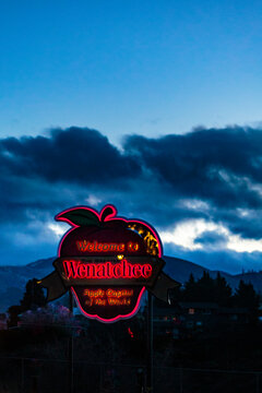 Welcome to Wenatchee town sign, Chelan County, an apple shape neon sign, under dark clouds in a blue sky.