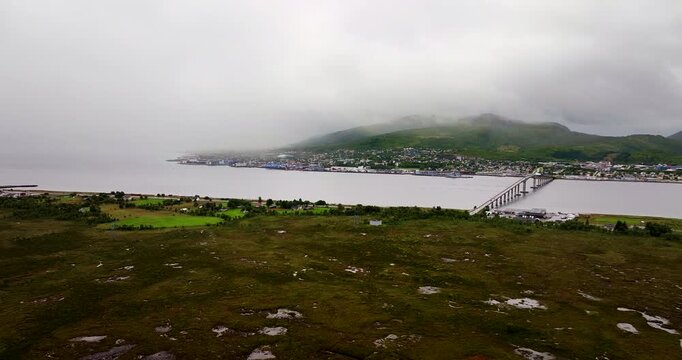 Wide aerial drone shot of Sortland in Vesteralen, Northern Norway. View of the bridge over the water with mist-covered mountain and a cloudy sky in the background