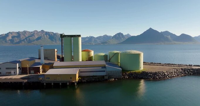 Drone flying over fish processing plant with silos on coast, with dramatic mountains of Lofoten Islands in background in Melbu, Norway. Aerial forward, copy space