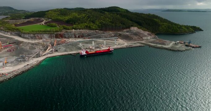 Red cargo ship being loaded with aggregate at port of open-pit stone quarry in Norway. Aerial drone, copy space