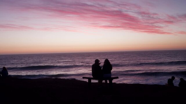 People sit in silhouette on a bench at Punta Lobos, Pichilemu, Chile. Peaceful view of the ocean during a pink sunset in South America. Perfect for travel and lifestyle content