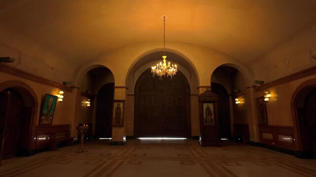 Expansive interior hall of Holy Trinity Cathedral, showcasing a grand chandelier illuminating the space. Features multiple arched doorways, a symmetrical layout, and large imposing wooden doors at ...