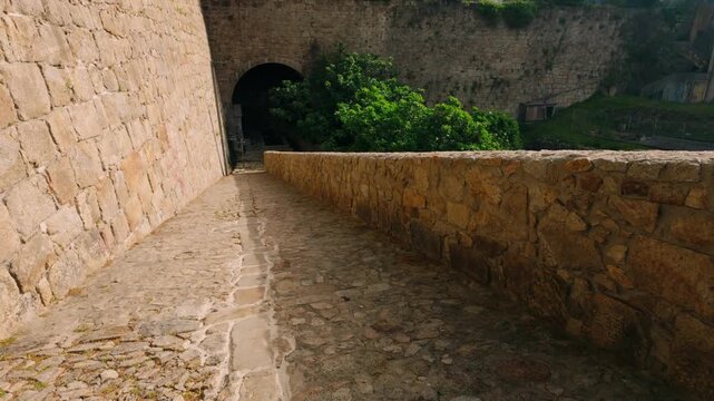 Historic cobblestone path winding alongside ancient stone walls, leading towards a dark, mysterious tunnel entrance. Lush green vegetation thrives along the weathered brickwork under bright natural...