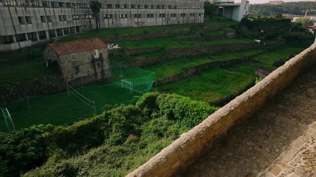 Elevated view showing terraced green fields with a traditional stone house and soccer goals. Modern buildings loom in the background, contrasting with the ancient stone wall in the foreground.