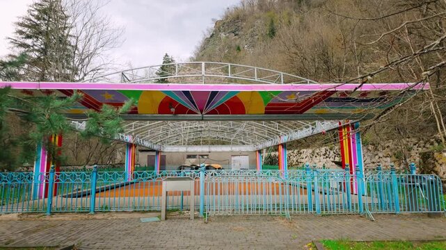 Empty bumper car ride area with a brightly colored canopy and railings in Borjomi Central Park. Forested mountains create a natural backdrop under a cloudy sky.