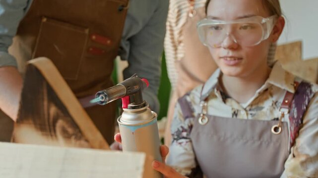 Rack focus view of teenage girl with safety glasses pointing propane torch at piece of wood while burning it at carpentry lesson in school, unknown teacher and classmate watching