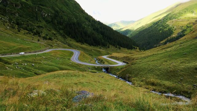 Cinematic panning shot of the winding Transfagarasan road in Romania. Scenic mountain valley with a small river and car driving at summer.