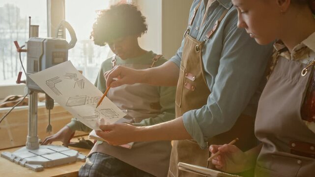 Unrecognisable male woodworker explaining paper sketch to two diverse teenagers taking notes during carpentry masterclass