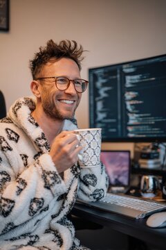 White male remote developer in pajamas sipping coffee and laughing at computer screen