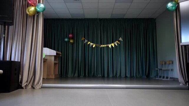 Assembly hall features curtains decorated with gold banner and balloons. Seating faces stage complemented by interactive display screen