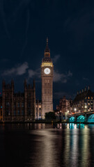 Obraz premium Bright clock face rises above the river and bridge at night, combining urban skyline with time concept and travel destination atmosphere.
