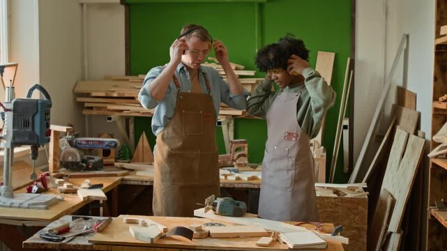 Caucasian gen Z carpenter and African American teenage apprentice putting on safety earmuffs and cutting wooden plank with power saw together