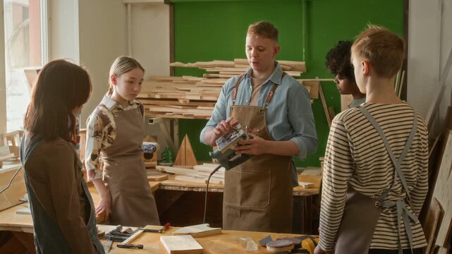 Gen Z male woodworker picking up jigsaw from table and explaining how to use it to group of four kids attending carpentry course