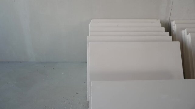 Minimalist composition of stacked white gypsum boards on gray concrete floor. Unfinished room at construction site with building materials ready for installation
