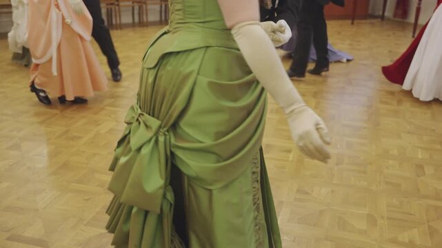 People in vintage costumes dance at a historical reenactment ball. Low angle shot follows couples waltzing on a wooden parquet floor