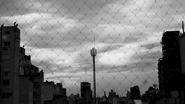 Black and white cityscape with chain link fence and cloudy sky