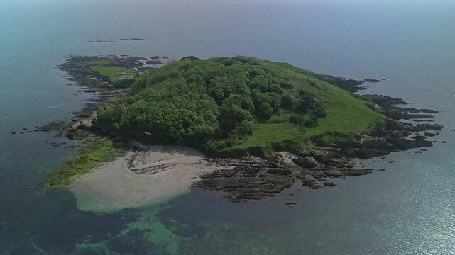 Cinematic 4K drone footage orbiting Looe Island in Cornwall, England. Bright sunshine illuminates calm turquoise water, rocky shoreline and lush island vegetation from an elevated aerial perspective.