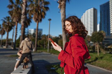Obraz premium Young woman with curly hair wears a red jacket while using her smartphone outdoors on a sunny day near palm trees and modern buildings.