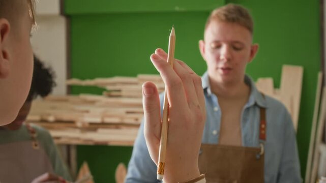 Over the shoulder rack focus shot of young woodworking instructor answering question of adolescent boy raising hand during explanation of how to work with bar clamp