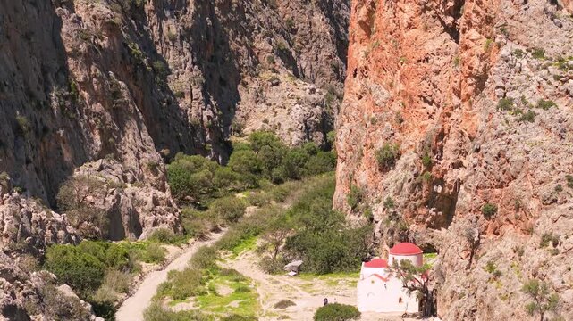 Aerial view of Agiofarango canyon church with red dome Crete Greece. Drone descending to Agios Antonios chapel and pull back reveal of steep orange cliffs in wild rocky gorge. Scenic landscape, 4k.