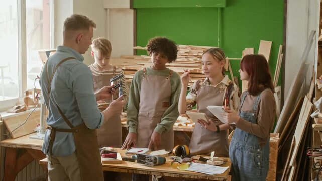 Young craftsman showing group of four teenage students how to use woodworking tools while they observing and taking notes in sunlit workshop