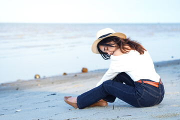 Young woman relaxing on a sandy beach, wearing a straw hat, and white sweater. She sits peacefully by the calm sea, enjoying a quiet moment of reflection. Perfect for themes of travel concepts.  © Said21 Photography 