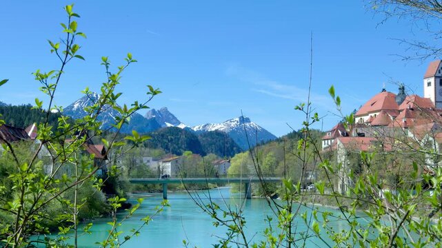 Tree branches swaying above the Lech River in F&uuml;ssen, Germany, with alpine mountains, turquoise water and soft focus spring landscape in sunny daylight