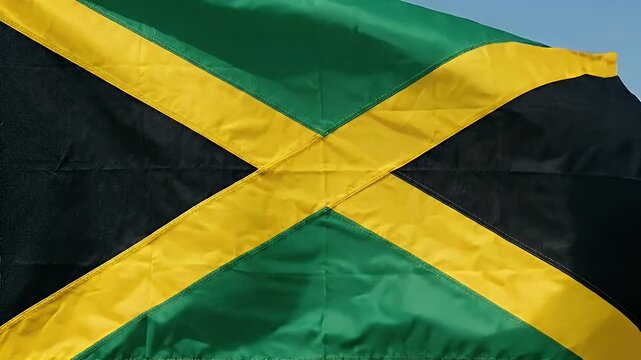 The video captures the vibrant Jamaican flag, prominently featuring green, black, and yellow colors. The flag is seen waving gracefully against a clear blue sky, symbolizing the rich cultural