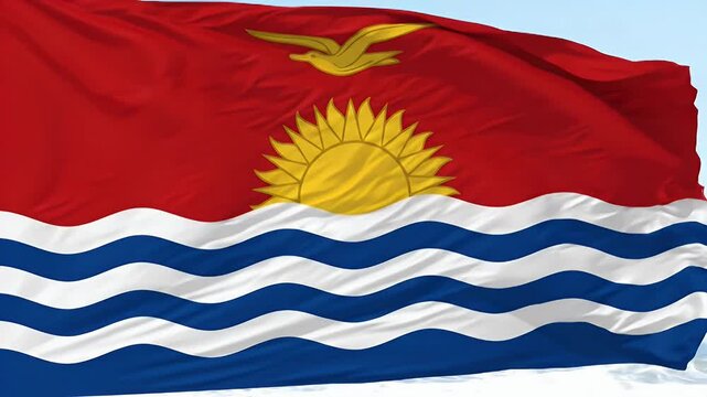 The video displays the national flag of Kiribati waving in the wind against a clear blue sky. The flag features a red background with a yellow frigatebird flying over a gold sun with seventeen rays,