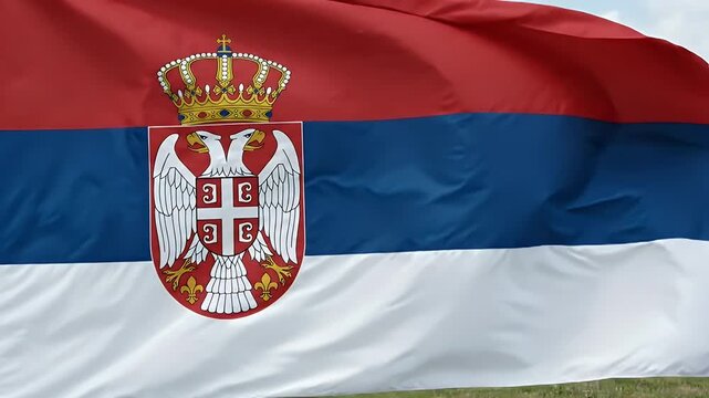 The video shows the national flag of Serbia waving gracefully against a clear sky. The flag features horizontal stripes of red, blue, and white, adorned with the Serbian coat of arms prominently