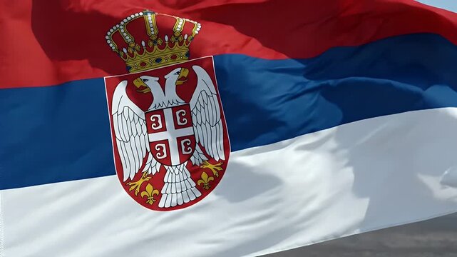 A vibrant Serbian flag waves in the wind against a clear blue sky. The flag features horizontal stripes of red, blue, and white, with a double-headed eagle emblem and crown in the center.