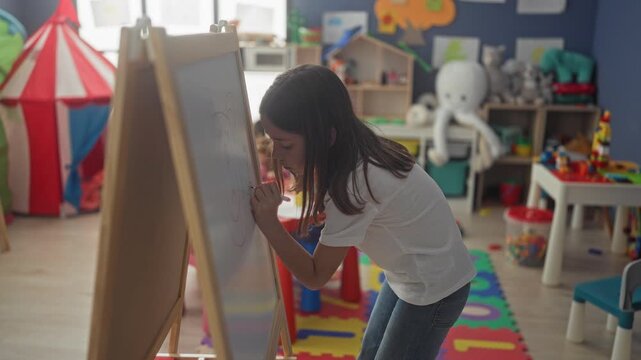 Girl drawing on easel with marker in colorful playroom, leaning close and holding marker to board; concentration imagination creativity.