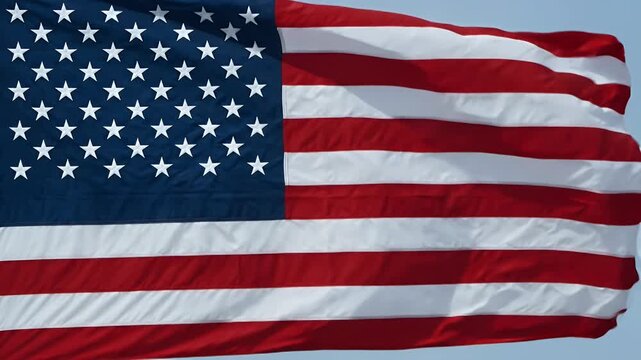 A close-up view of the United States flag gently waving in the breeze. The flag features its iconic red and white stripes with a blue field adorned with white stars, symbolizing the nation&rsquo;s unity