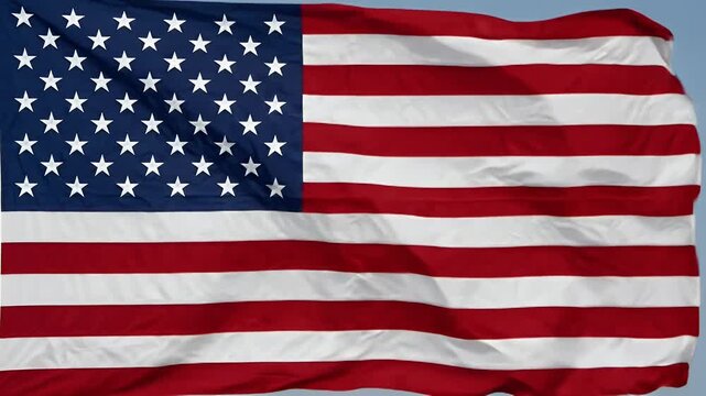 The video shows the flag of the United States billowing against a clear blue sky. The flag features its iconic design of 50 white stars on a blue field and 13 alternating red and white stripes,