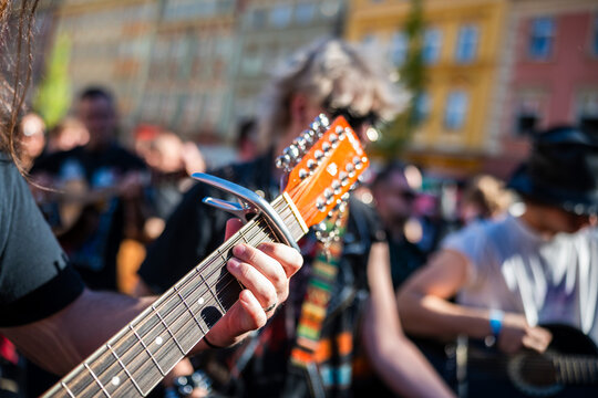 Close-up shot of a musician playing a 12-string acoustic guitar with a capo during an outdoor street performance, focusing on the left hand and the headstock.