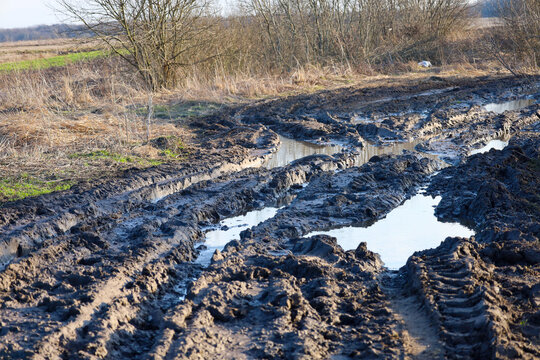 ​A rural dirt road is completely washed out by the spring thaw and covered with deep muddy ruts and large puddles. An impassable section of the path runs through a field near bushes,