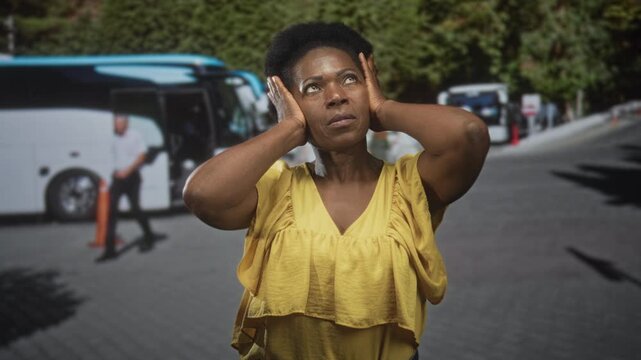 Woman covers ears with both hands beside parked coach buses on a street shuttle pickup, wearing yellow blouse and squinting in daylight; travel noise stress.