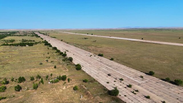 Aerial view of a lonely white car driving on a massive abandoned military runway overgrown with grass. Former secret Chagan airbase, Semipalatinsk Test Site, Kazakhstan. Cinematic post-apocalyptic lan