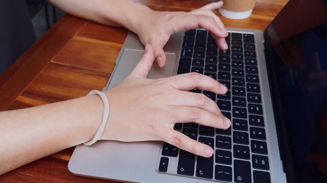 Person's hands typing swiftly on a laptop keyboard, using technology to connect, work, and manage business tasks remotely from a wooden table, with a cup of coffee nearby