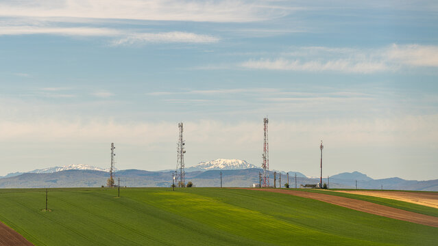View of telecommunication towers standing on rolling green agricultural fields with snow-capped mountains in the distance under a blue sky.