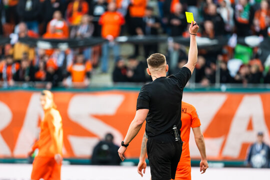 Back view of a soccer referee in a black uniform holding up a yellow card for a foul during a professional match in a crowded stadium.