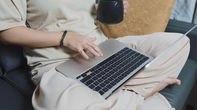 Woman multitasking on a laptop computer while holding a mug, sitting comfortably on a sofa, performing remote work and enjoying a relaxed home office lifestyle
