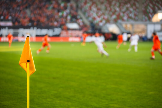 A close-up of a bright yellow corner flag on a grass pitch with a football match and stadium crowd blurred in the background.