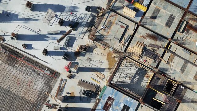 Top-down aerial view of a large construction site in its early stages, showing concrete foundations, steel reinforcing bars, and wooden formwork under bright sunlight casting long shadows