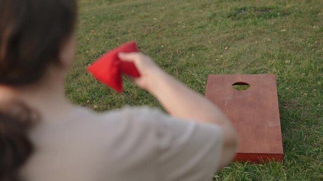 Woman preparing to throw cornhole bag, squeezing and tossing equipment in hands, view over the shoulder. Player throws red bean bag, it lands on wooden board surface near hole.