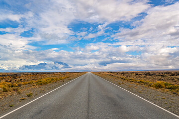 Fototapeta premium Straight asphalt road leading towards distant mountains under a blue sky with dramatic clouds in Patagonia landscape