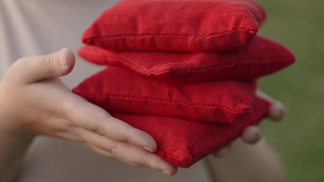 Closeup of set of red bean bags sized 6 inches for cornhole game in hands. Unrecognizable player holding set of tossing equipment with slow slide speed.